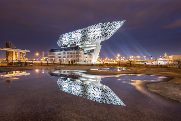 Nachtelijke scene met het Zaha Hadid Havenhuis Antwerpen gereflecteerd in een waterplas. 